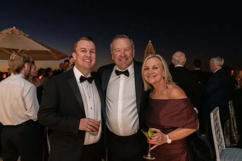 Craig stands between two men in tuxedos while a woman in a burgundy dress smiles nearby during an outdoor evening reception.