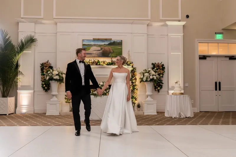 Ashley and Craig walk hand in hand across the ballroom beneath floral garland.