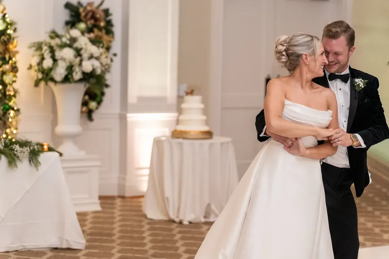 Ashley and Craig embrace in the ballroom, with the wedding cake visible on a draped table behind them.