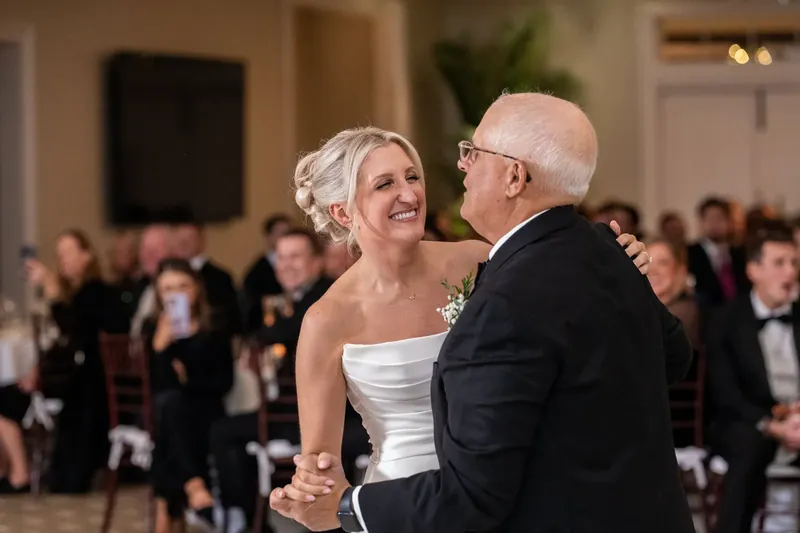Ashley dances with an older man in a dark suit, both smiling as reception guests watch from their seats.