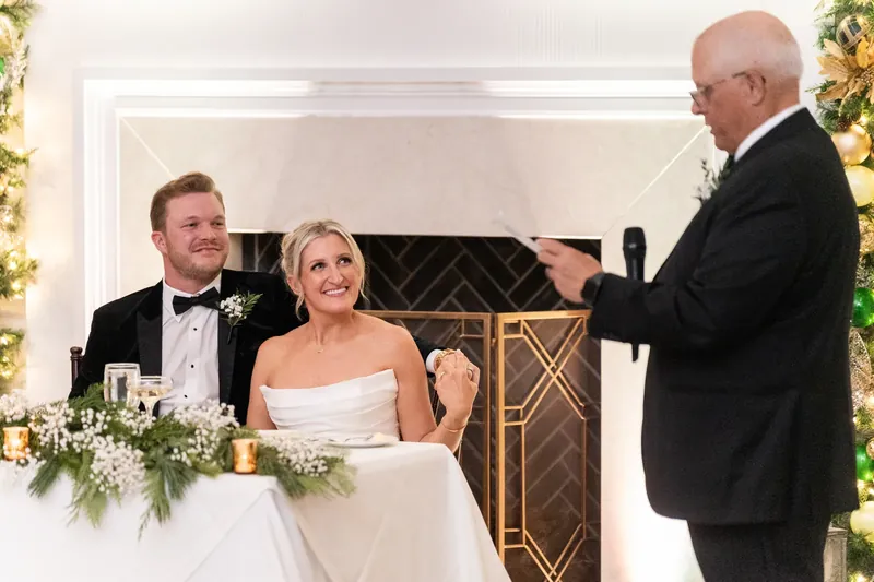 Craig and Ashley sit at their table while a man in a tuxedo speaks and gestures beside them.