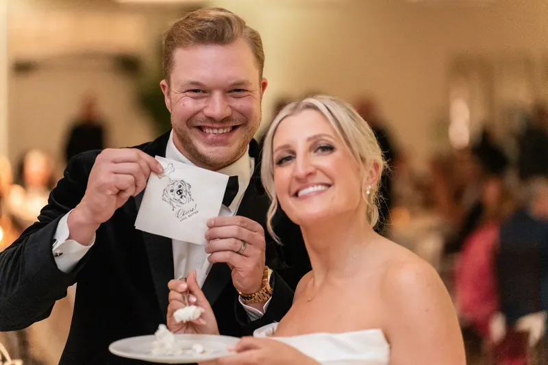 Craig and Ashley laugh while holding a napkin with a bite of cake, their joy evident as guests celebrate behind them.
