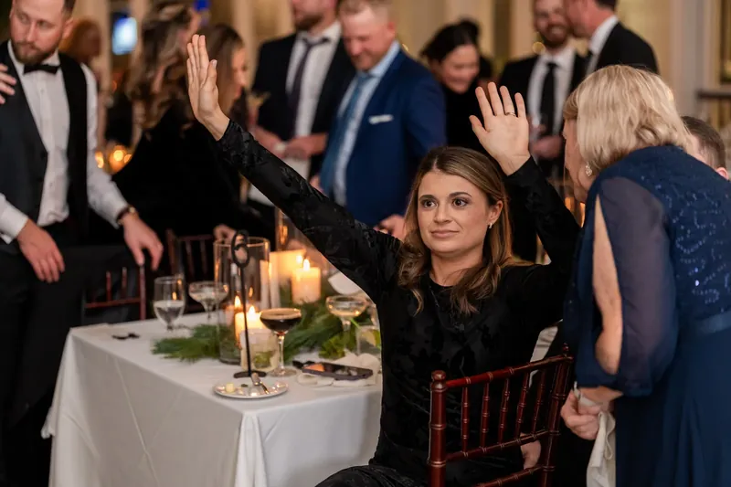 Ashley waves from her seat at a banquet table while wedding guests around her raise their hands in celebration.