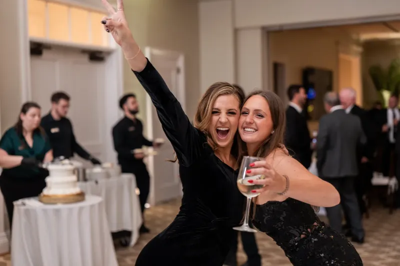 Two women raise their champagne glasses and pump their fists in the air while dancing at the reception.