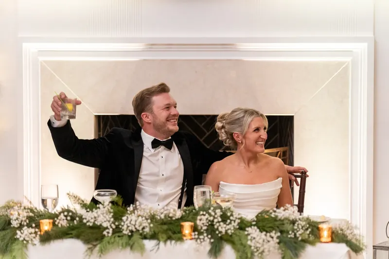 Craig raises his fist in triumph at the head table, seated next to Ashley, with greenery and candles adorning the space.