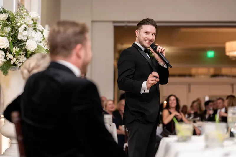 Craig speaks into a microphone at the reception while another guest listens from nearby.