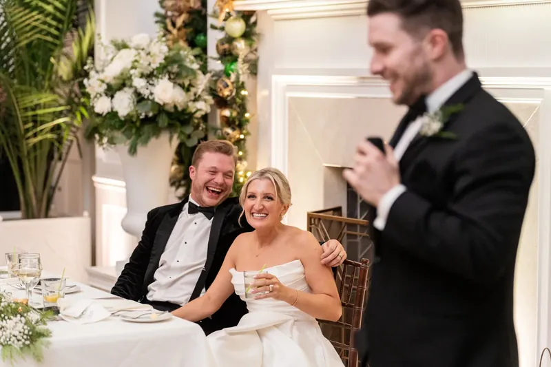 Craig listens as a groomsman speaks during toasts at the reception, with Ashley smiling beside him at their table.