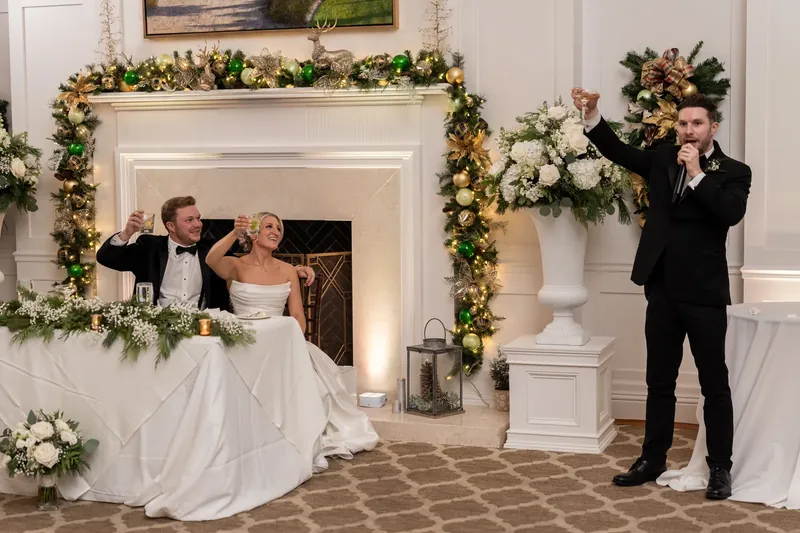 Ashley and Craig sit at their sweetheart table by a fireplace draped with garland while someone speaks during the reception.
