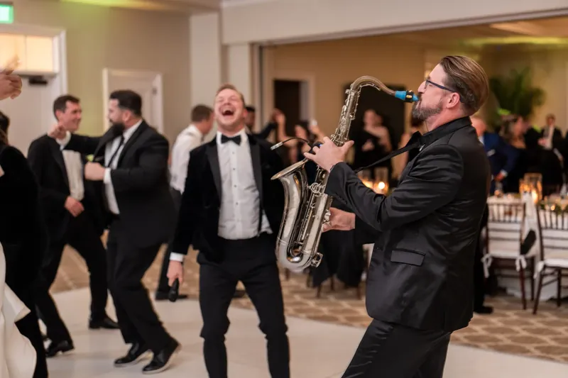 A saxophonist in black tie plays energetically as wedding guests dance and mingle in the reception space.