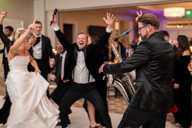 Ashley dances in her white gown alongside a saxophonist and guests in formal attire during the reception.