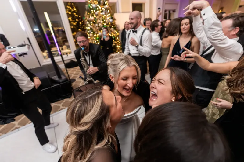 Two women laugh together at the reception, with partygoers and holiday garland visible throughout the festive ballroom.