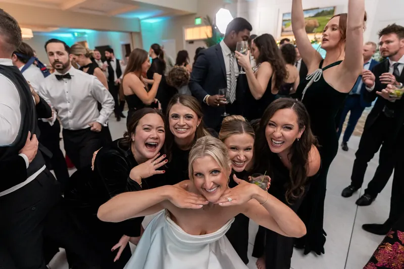 Ashley sits surrounded by smiling friends in black attire, arms around each other in the reception hall.
