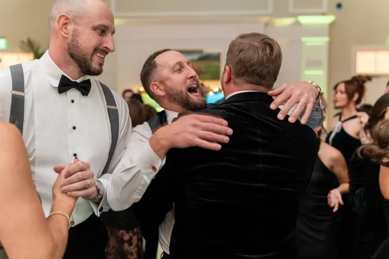 Three men in formal wear embrace and laugh together during the reception.