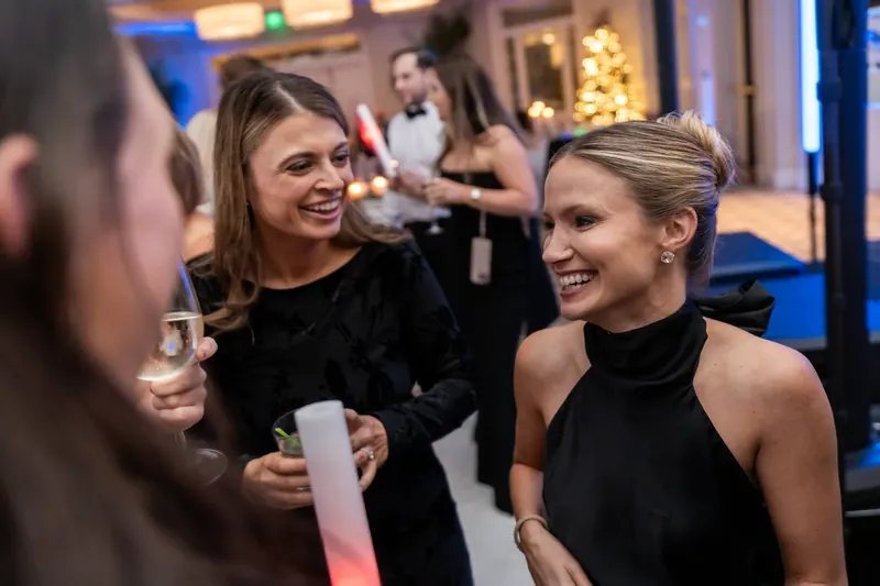 Two women in black dresses smile while chatting and holding drinks at the celebration.