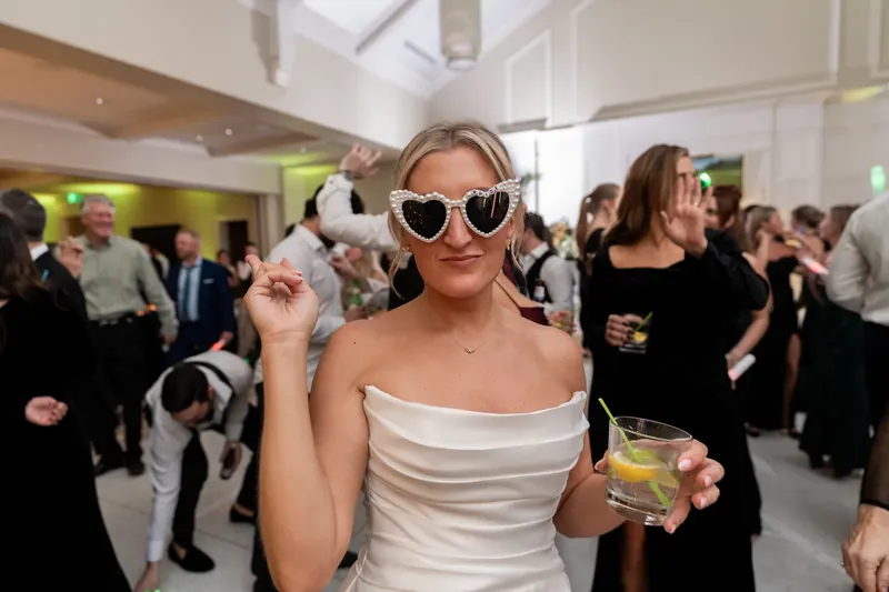 A woman in a white off-shoulder dress wears oversized sunglasses and holds a drink while dancing.