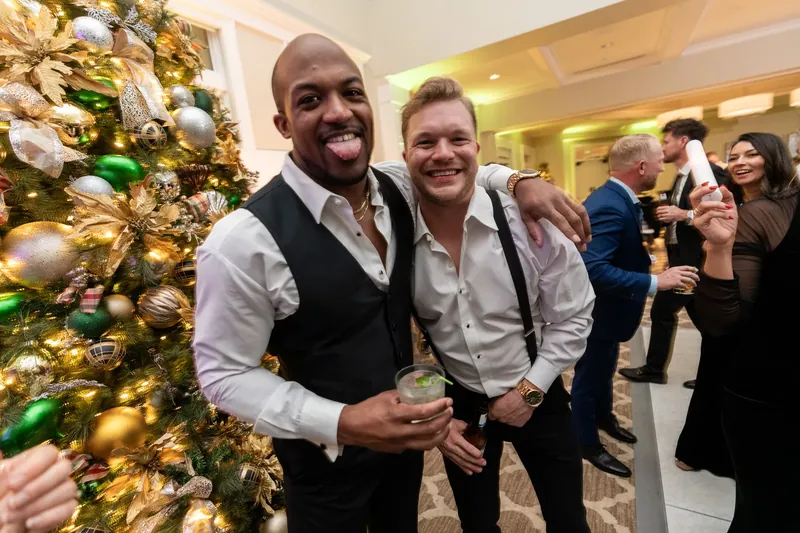 Two men in formal wear smile beside a decorated garland at the Sawgrass Marriott reception.