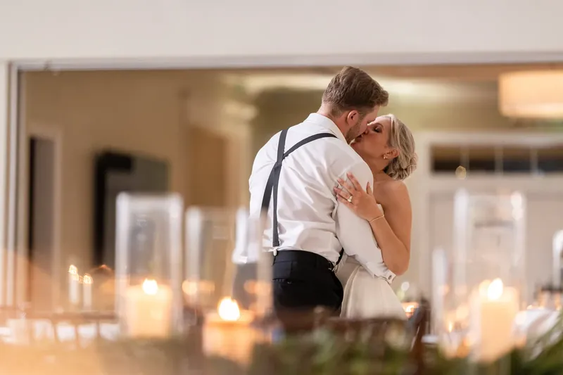 Craig leans down to kiss Ashley's cheek as candles flicker softly in the foreground of the elegant reception space.