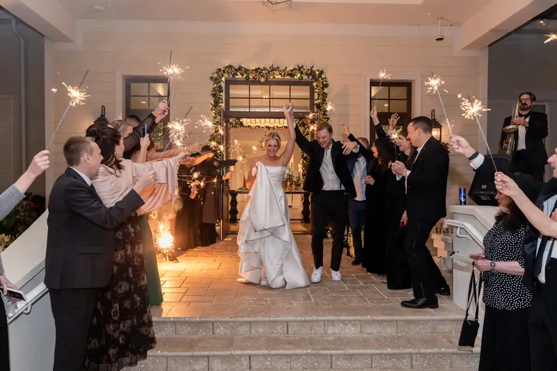 Ashley and Craig walk through a wreath-framed doorway as guests hold up sparklers on either side.