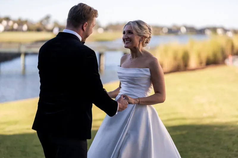 Craig holds Ashley's hand as they face each other, smiling, with a waterway and golden marsh grasses stretching behind them.