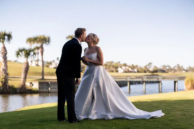 Craig kisses Ashley's forehead as she holds her white and green bouquet, their silhouettes framed by palm trees and water.