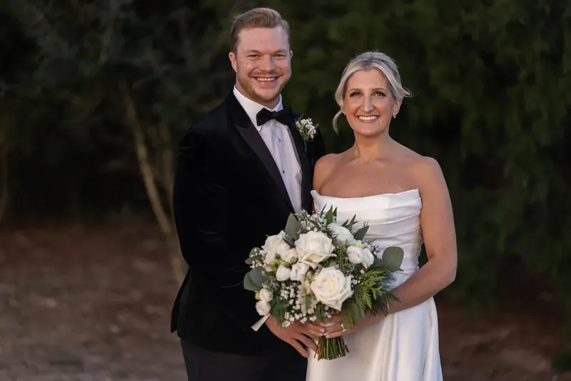Craig and Ashley stand together at dusk, both smiling at the camera, with Ashley holding a white and green bouquet.