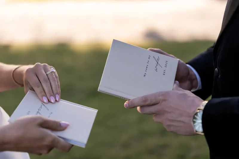 Two hands exchange white ceremony programs during golden-hour light with a lawn visible beyond.