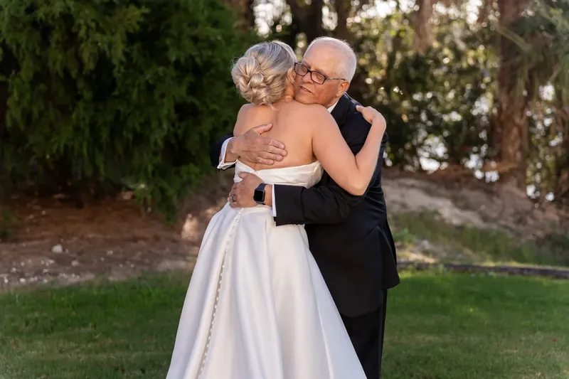 Ashley embraces an older man in glasses while wearing her white wedding dress on the grass.