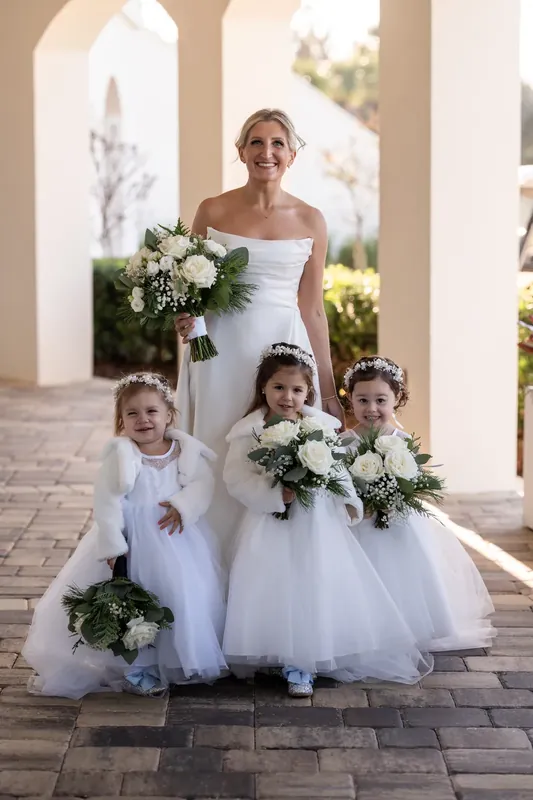 Ashley holds a cascading bouquet while posing with three flower girls in white dresses on a covered patio.