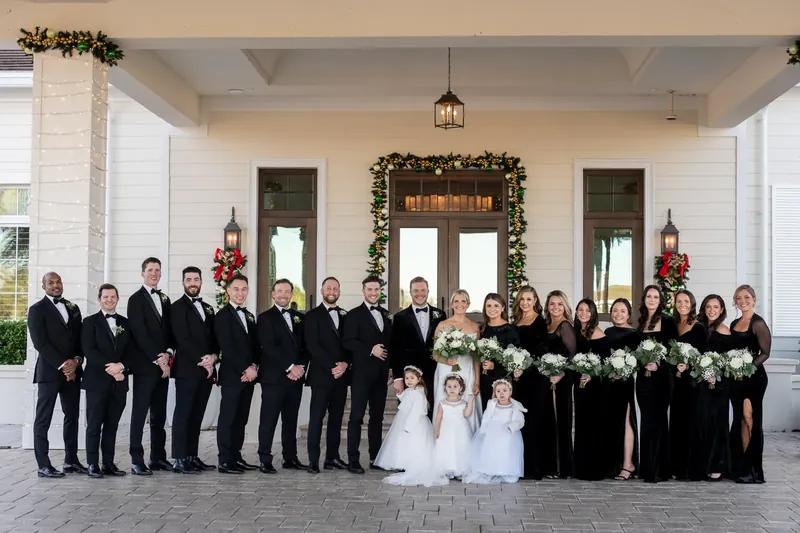 Ashley and Craig pose with their entire wedding party on the columned porch, holding white florals.