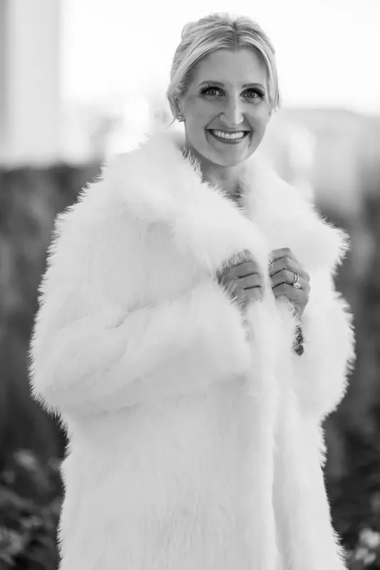 Ashley wears a white fur stole over her gown, smiling warmly at the camera outdoors.