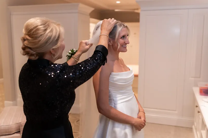 A woman adjusts Ashley's hair in an upswept style while Ashley stands in her white wedding dress in an indoor bridal suite.