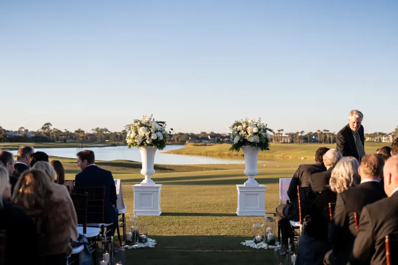 Guests seated on the lawn watch the ceremony setup with white pedestals of florals across the golf course.