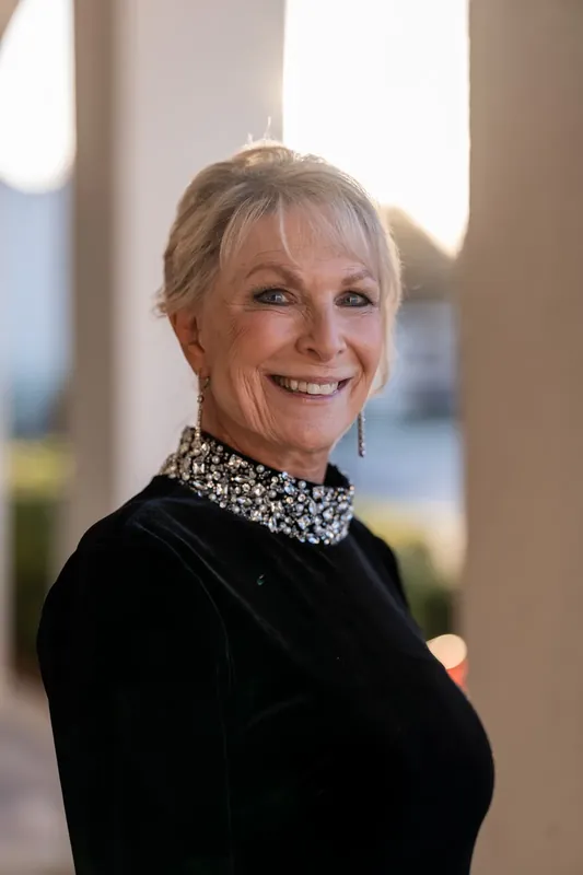 A woman in a black sequined jacket and beaded collar smiles at the camera with natural light illuminating her face.