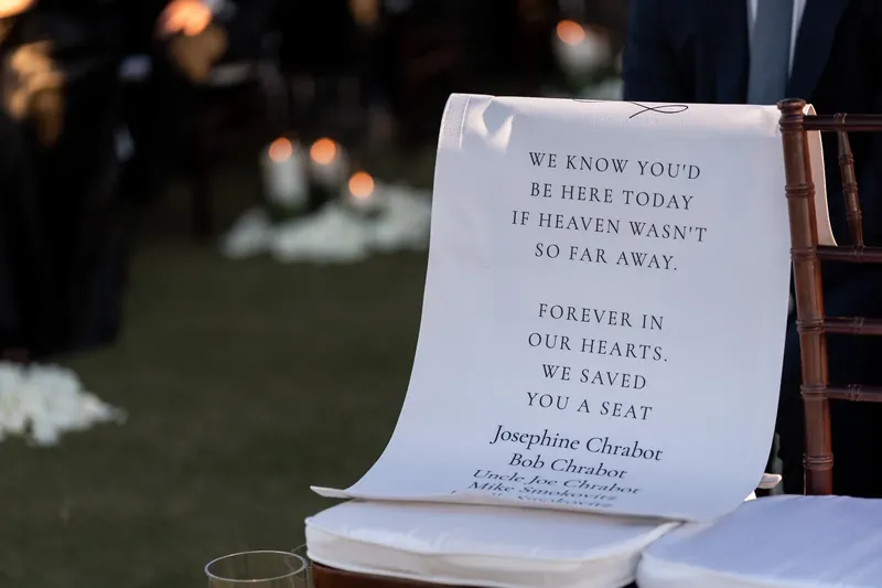 A memorial card reading "We know you'd be here today" rests on a white ceremony chair, honoring absent loved ones.