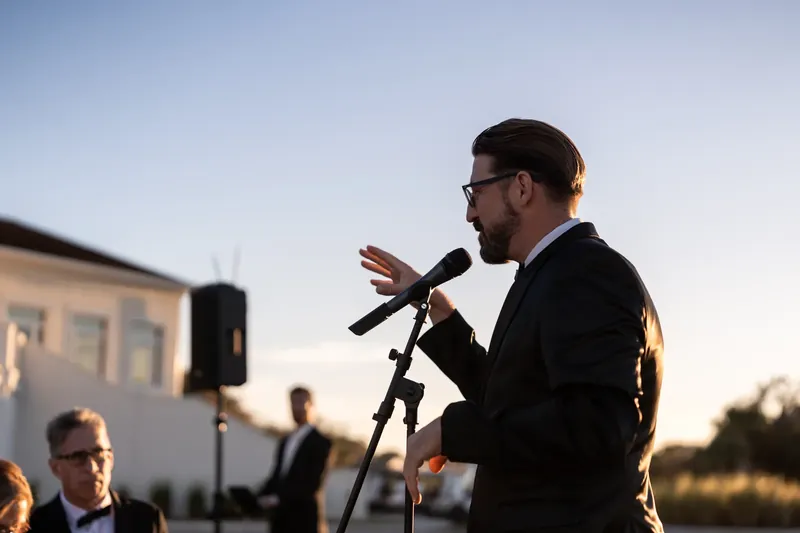 Craig speaks at the microphone during the outdoor ceremony as guests listen along the fairway at dusk.