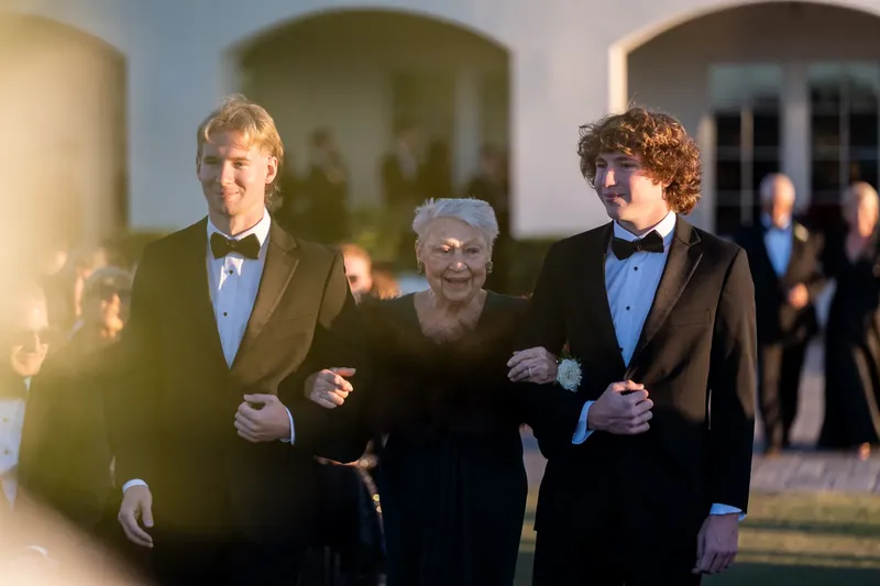 Two groomsmen escort an older family member down the aisle past the arched colonnade.