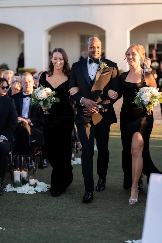 Ashley walks arm-in-arm with two attendants, holding a white bouquet, in her entrance down the aisle.
