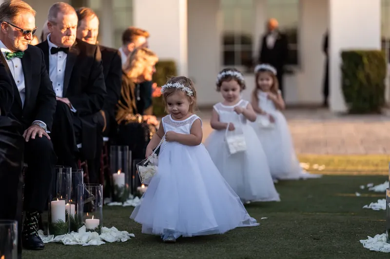 Young flower girls in white dresses stand on the grass during the ceremony as guests watch.