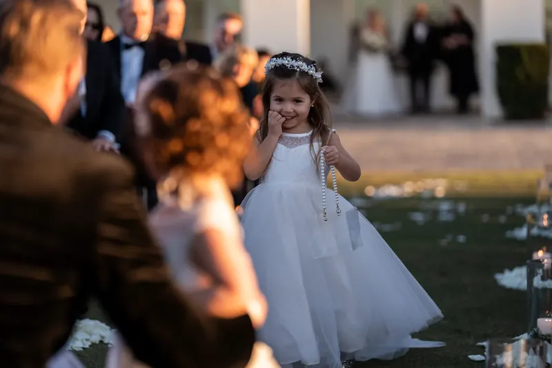 A flower girl in a white dress smiles while standing near the aisle during the ceremony.