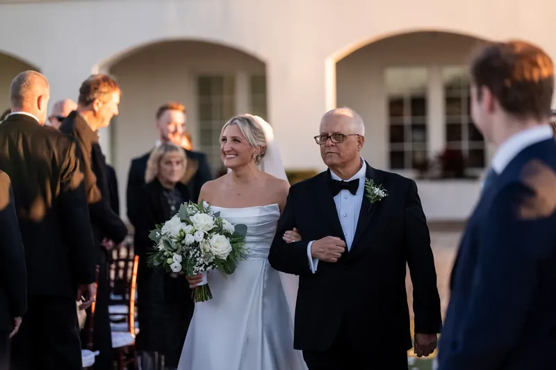 Ashley, wearing a white gown and veil, walks arm-in-arm with a family member past guests near the resort's building.