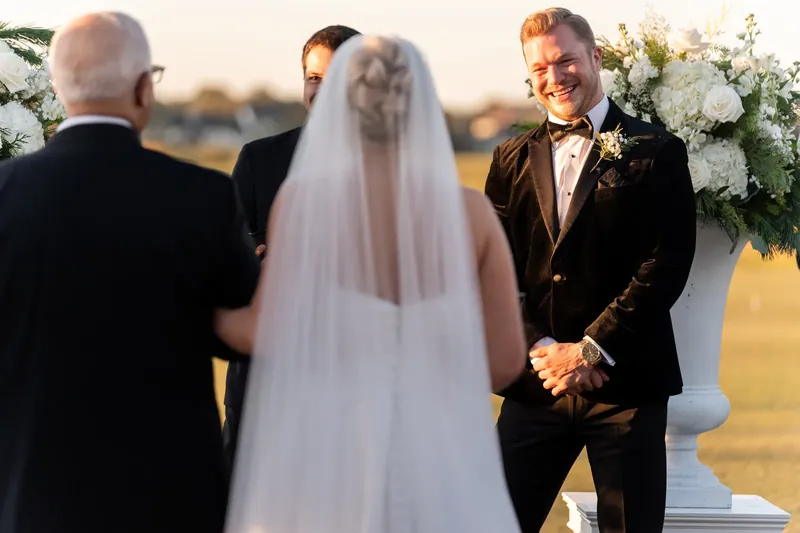 Craig and Ashley stand at the altar as an officiant presides over their ceremony with white floral arrangements nearby.