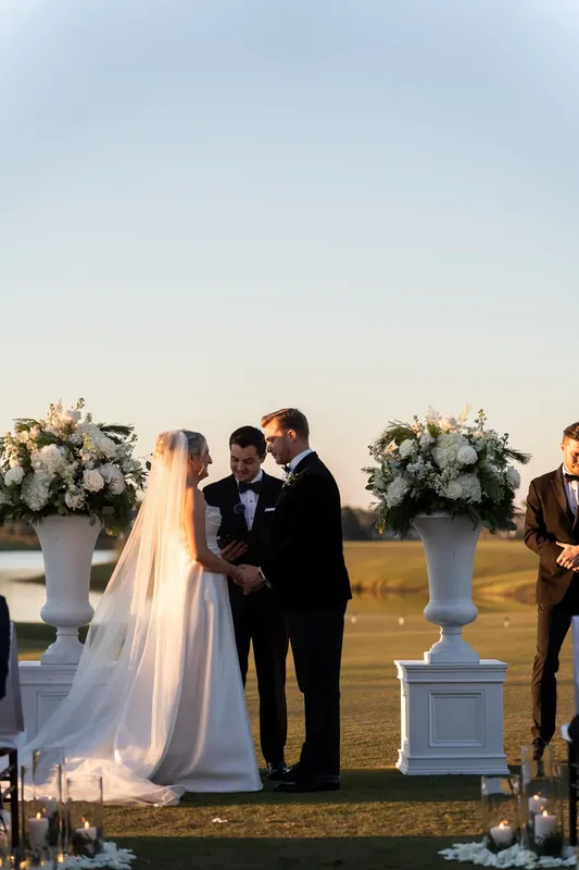 Ashley and Craig face each other at the altar with white florals and water behind them at sunset.