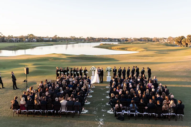 Overhead view of the wedding party and guests arranged on the manicured golf course by the water.