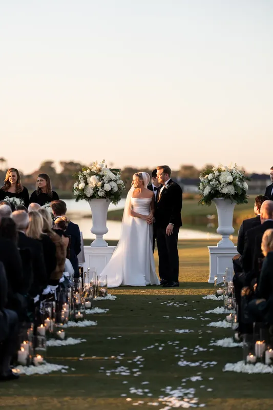 Ashley and Craig walk down the flower-strewn aisle as standing guests applaud between white arrangements.
