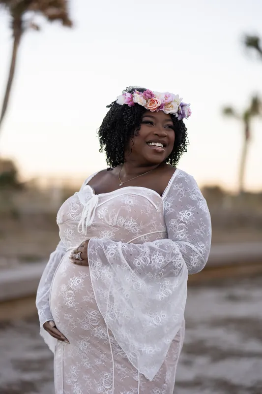 Ashley smiles at the camera while holding her baby bump, wearing a white lace dress and pink floral crown against