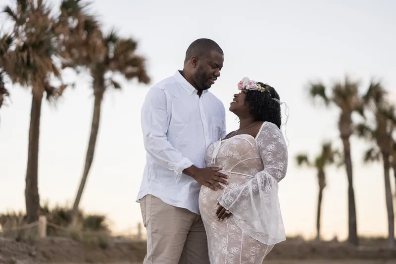 JB gently touches Ashley's pregnant belly while standing close together on the beach, with tall palms visible behind them