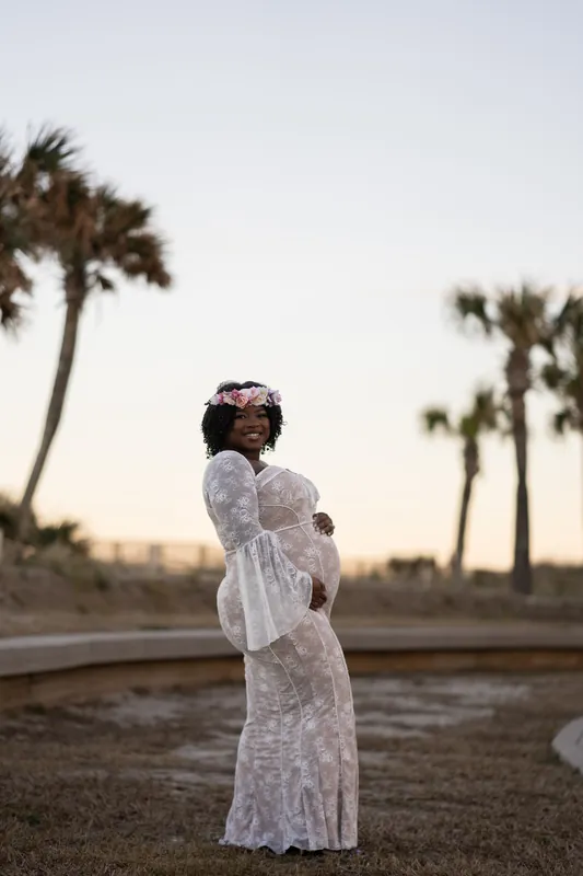 Ashley poses in profile on the beach, one hand on her belly, wearing the sheer white lace maternity dress with palm trees