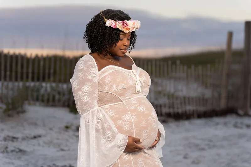 Cradling her baby bump, Ashley wears a lace maternity dress and flower crown near coastal dunes at sunset.
