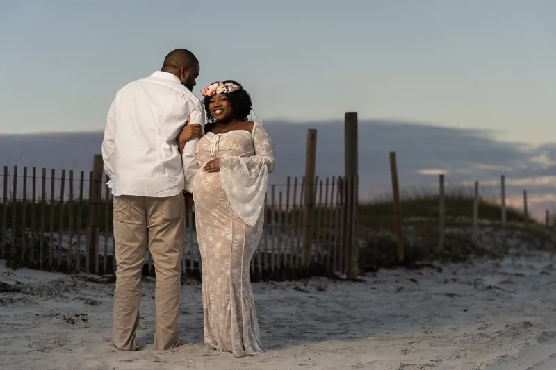 JB wraps his arms around Ashley from behind as they stand on the beach with a wooden fence visible behind them.