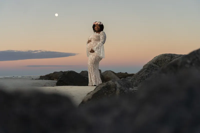 Ashley stands alone on a rock outcropping in her lace dress, silhouetted against the twilight sky at the beach.